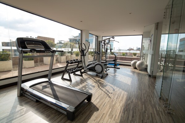 A modern gym with a treadmill, elliptical, and weight machines, large windows with a city view, and a wooden floor.