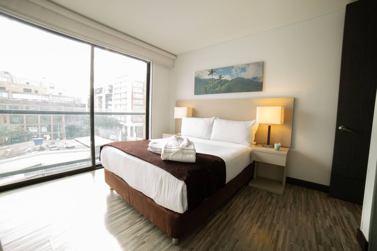 A hotel room with a large window, a bed with white linens, a brown headboard, a nightstand with a lamp, and a painting on the wall.