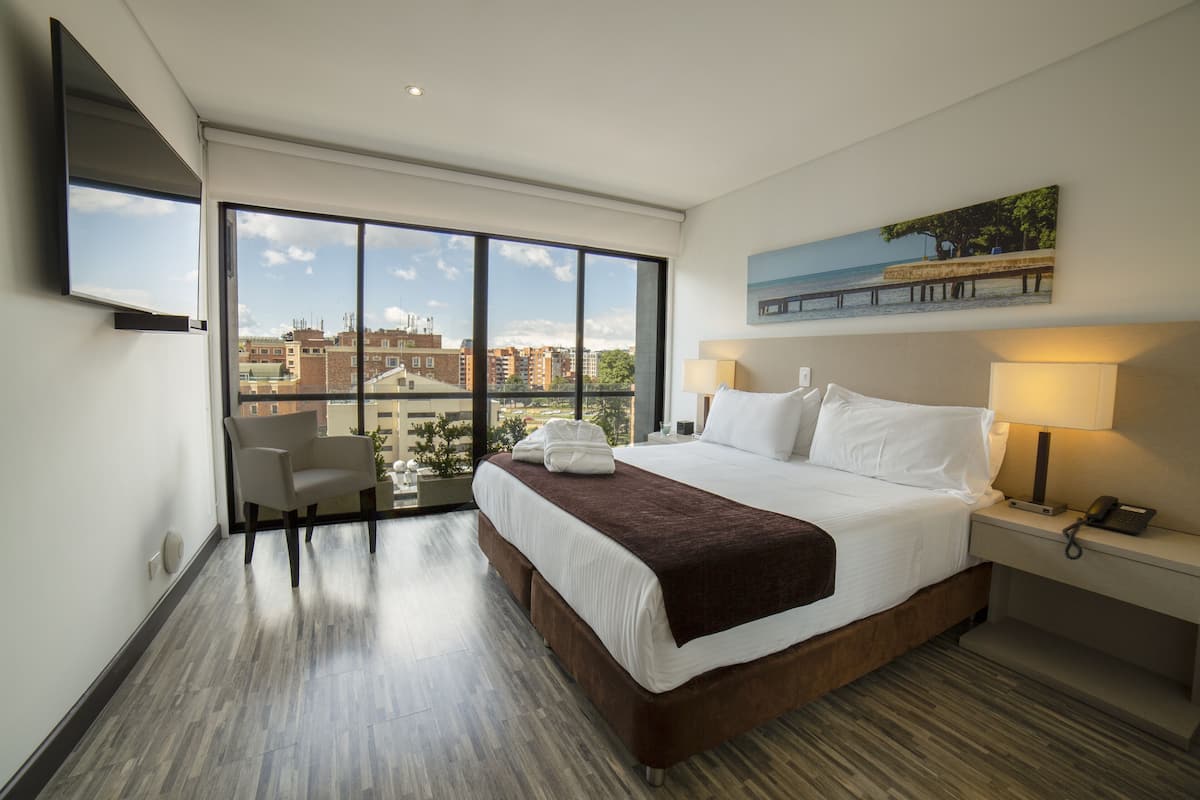 A hotel room with a large bed, a chair, a desk with a lamp, and a painting on the wall.