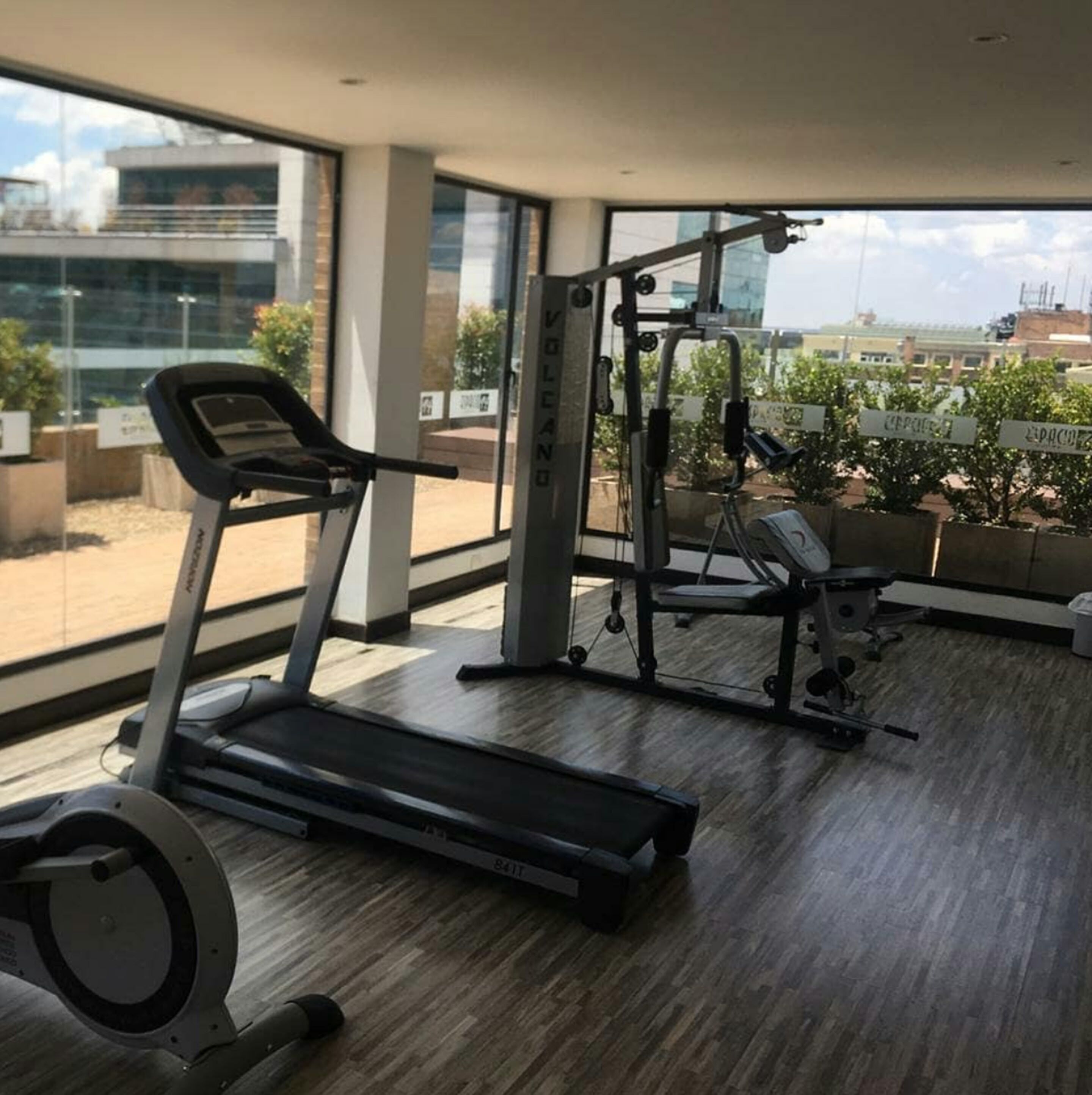 Une salle de sport moderne équipée de tapis de course, d’appareils de musculation et d’un banc. De grandes fenêtres offrent une vue sur la ville.