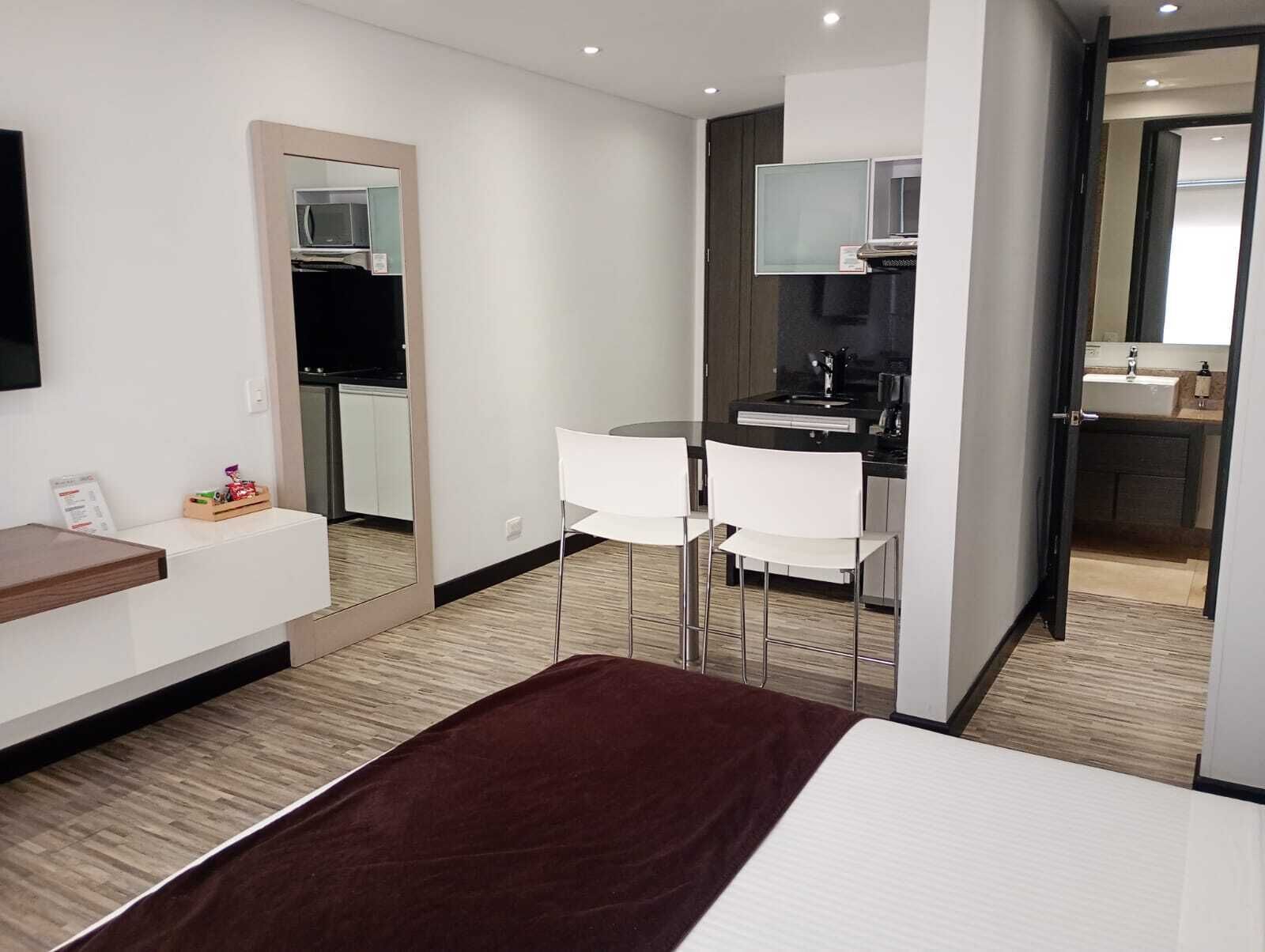 A modern hotel room with a bed, a small desk, a mirror, a kitchenette, and a bathroom.