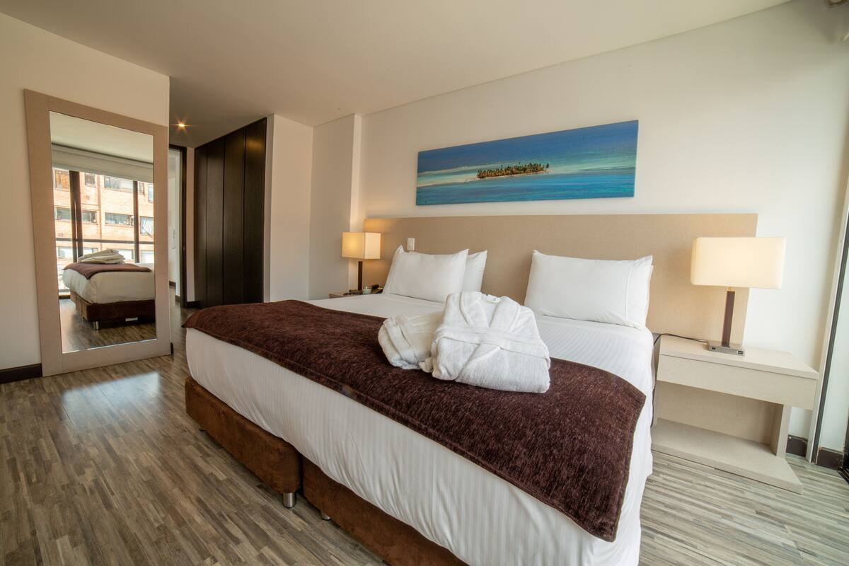 A hotel room with a large bed, two bedside tables, a painting on the wall, and a view of the outside through a door.