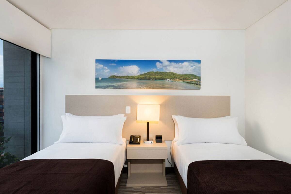A hotel room with two beds, a nightstand with a lamp, a painting of a beach scene, and a window with a view of a building.