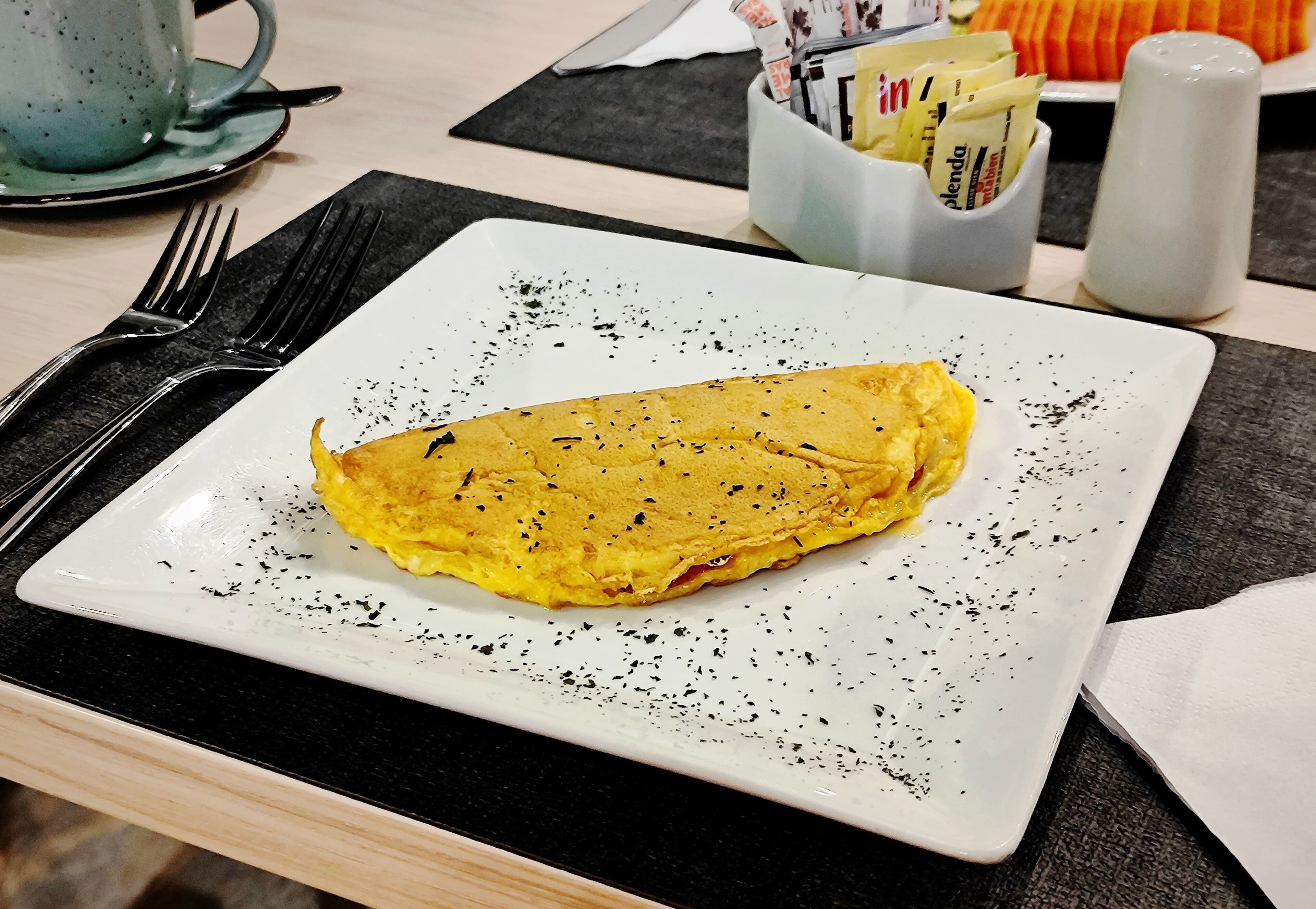 A plate with an omelette on a white square plate, accompanied by a fork and a salt shaker.