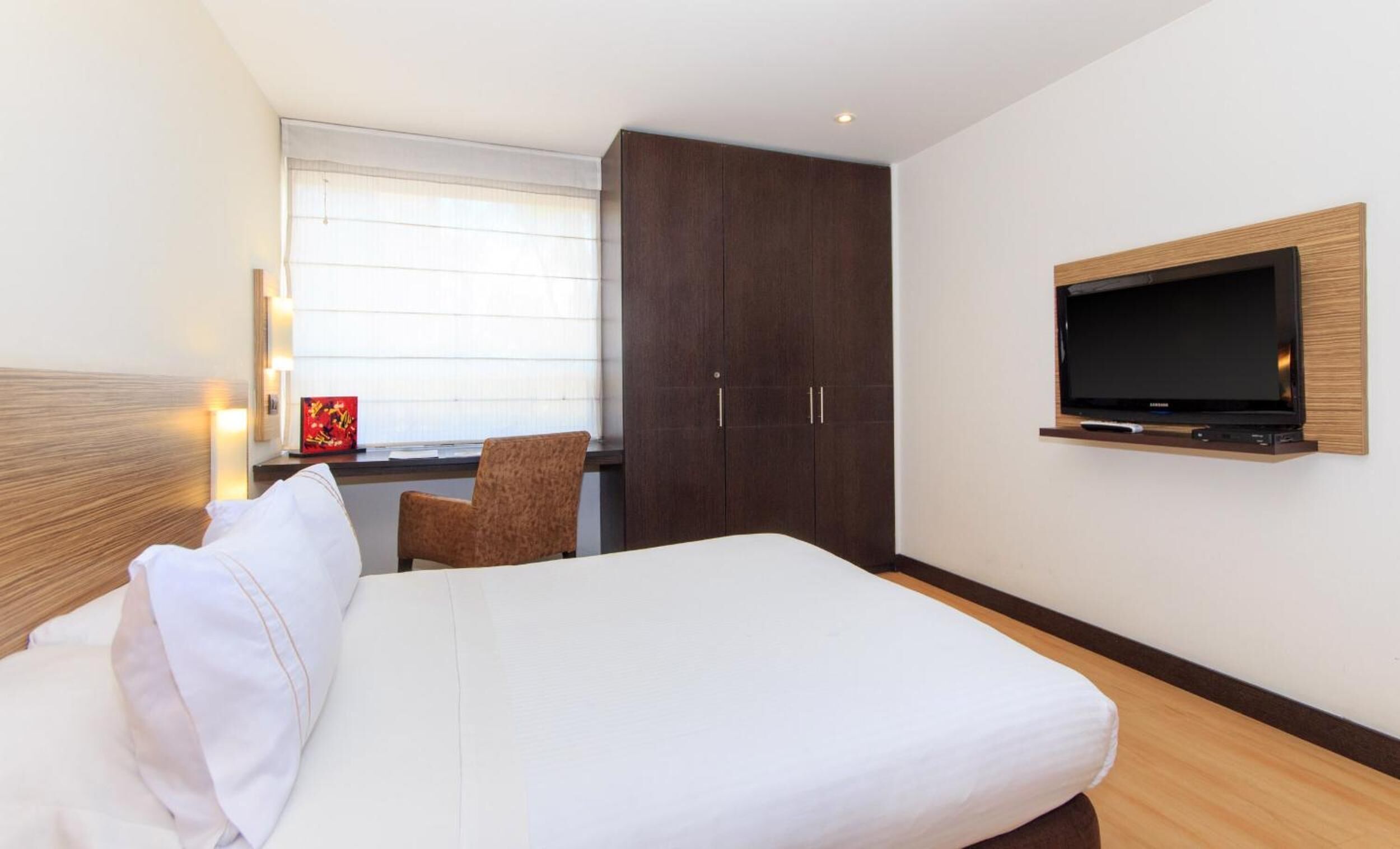 A hotel room with a bed, a TV mounted on the wall, a wardrobe, and a window with blinds.