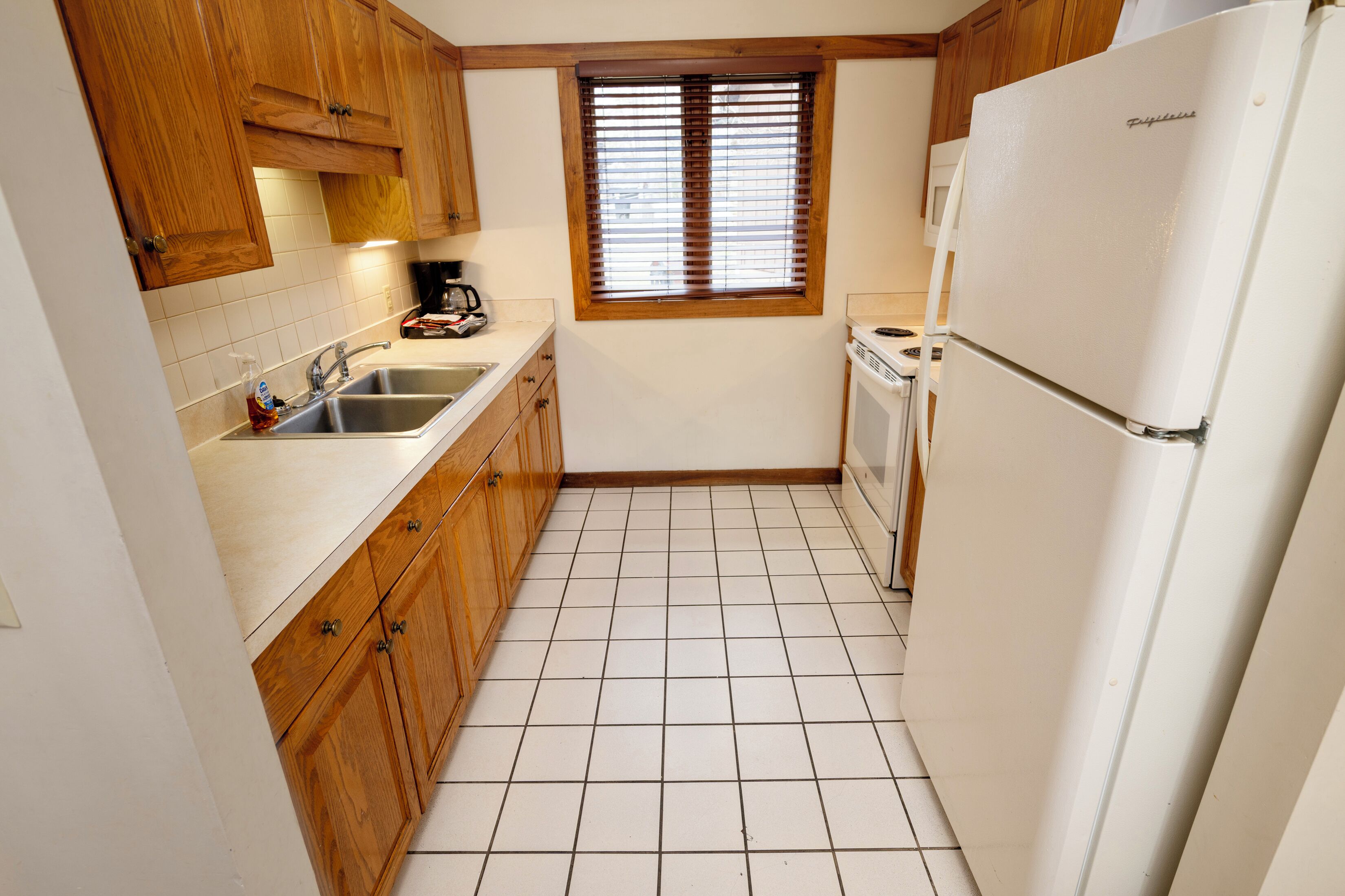 A compact kitchen with wooden cabinets, a white refrigerator, and a window with blinds.