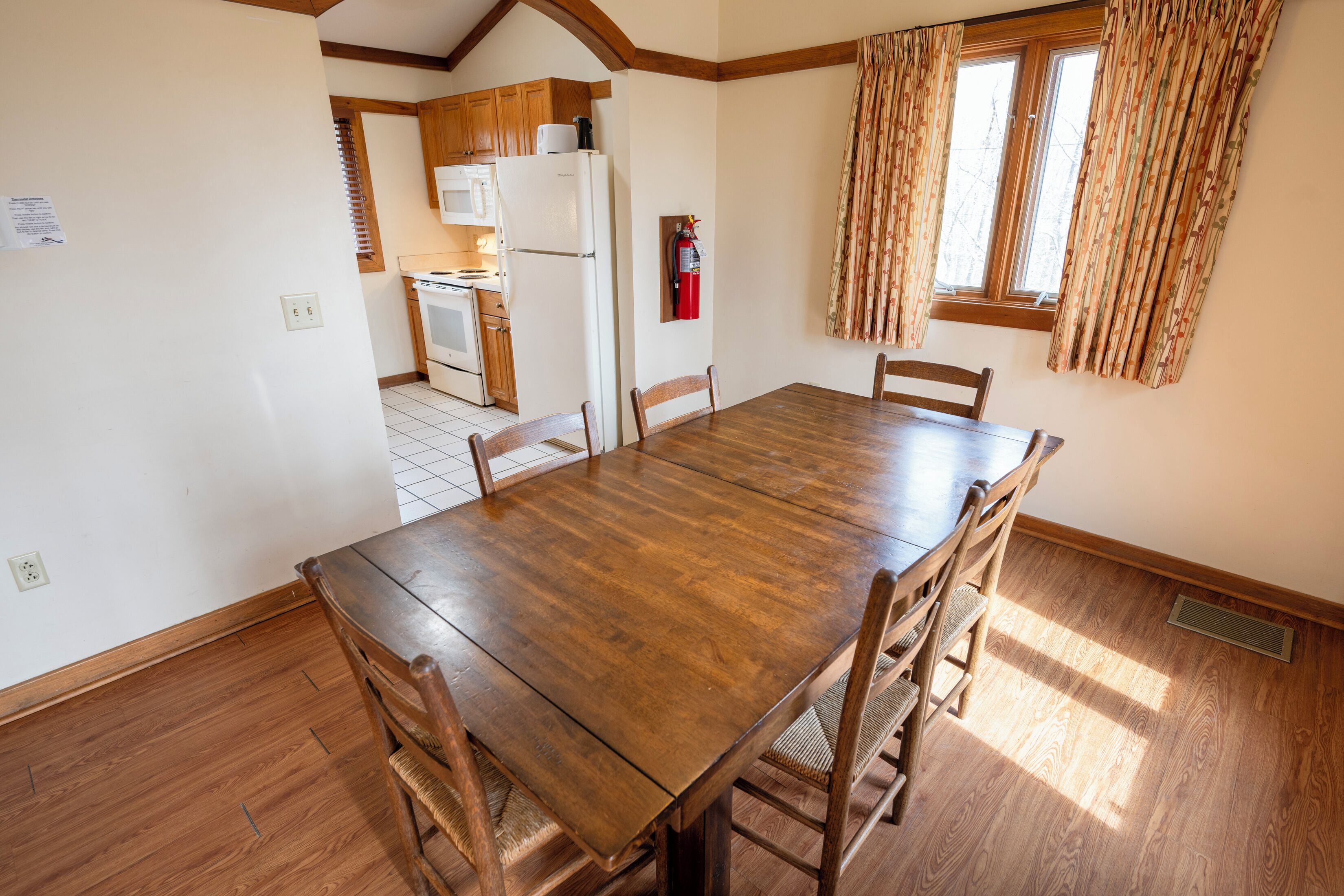 Un coin repas avec une table et des chaises en bois, une cuisine à l’arrière-plan et une fenêtre avec des rideaux.