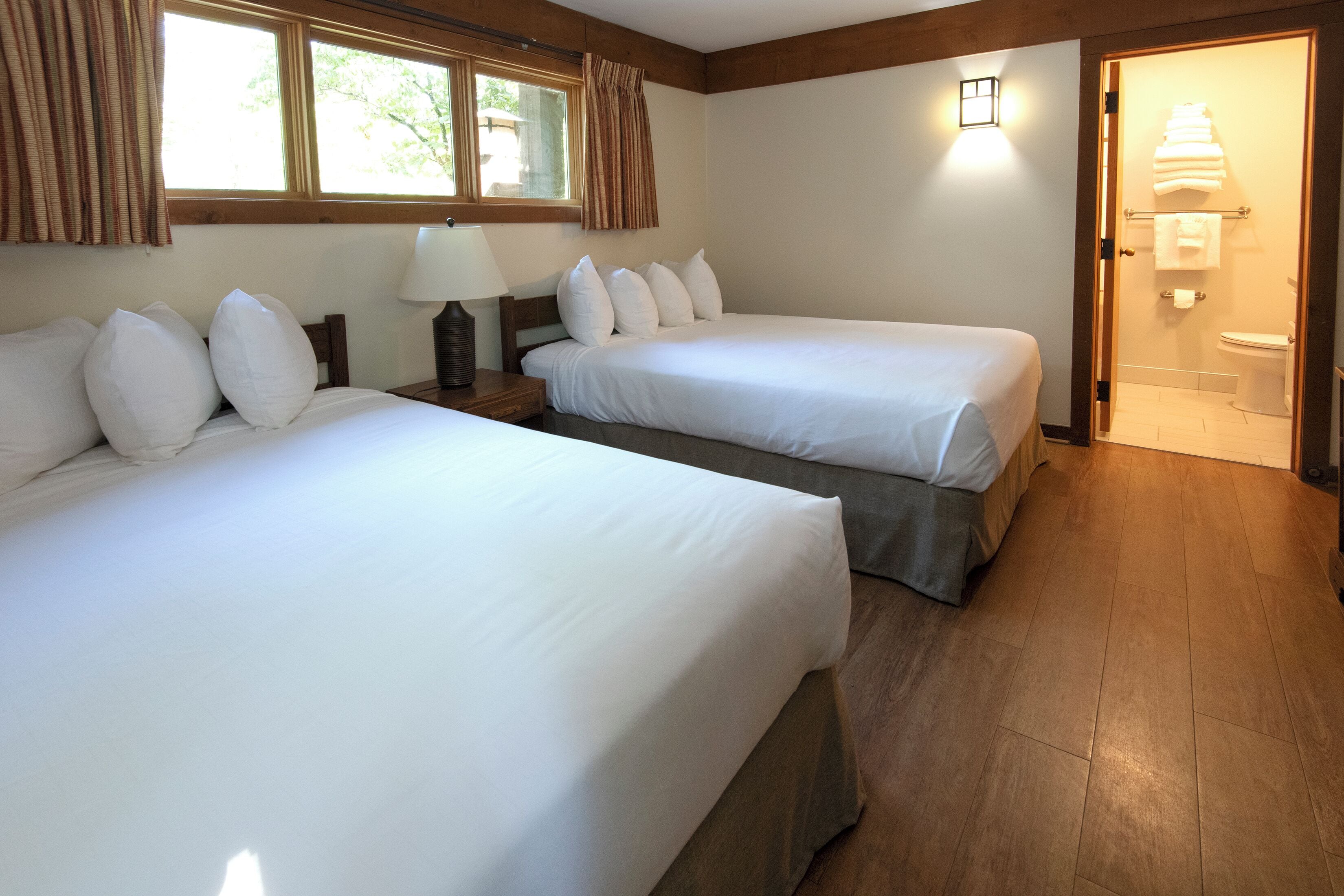 Habitación de hotel con dos camas individuales, una mesita de noche con lámpara, una ventana con cortinas, suelo de madera y un baño con toallero.