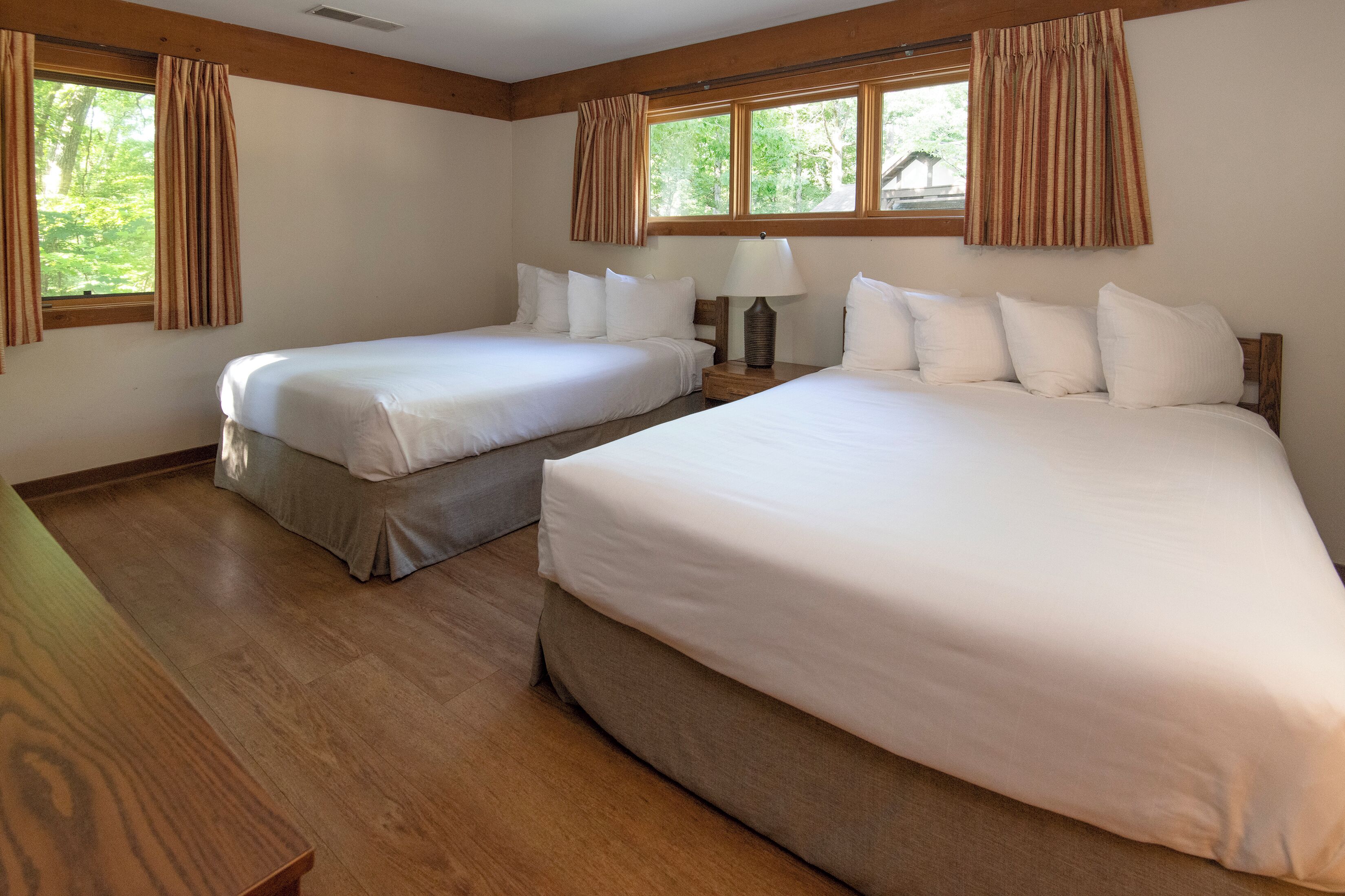 Quarto de hotel com duas camas, piso de madeira e grandes janelas com cortinas.