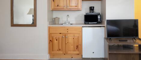Een kleine keuken met houten kastjes, een magnetron, een spoelbak en een ingebouwde televisie op een houten standaard.