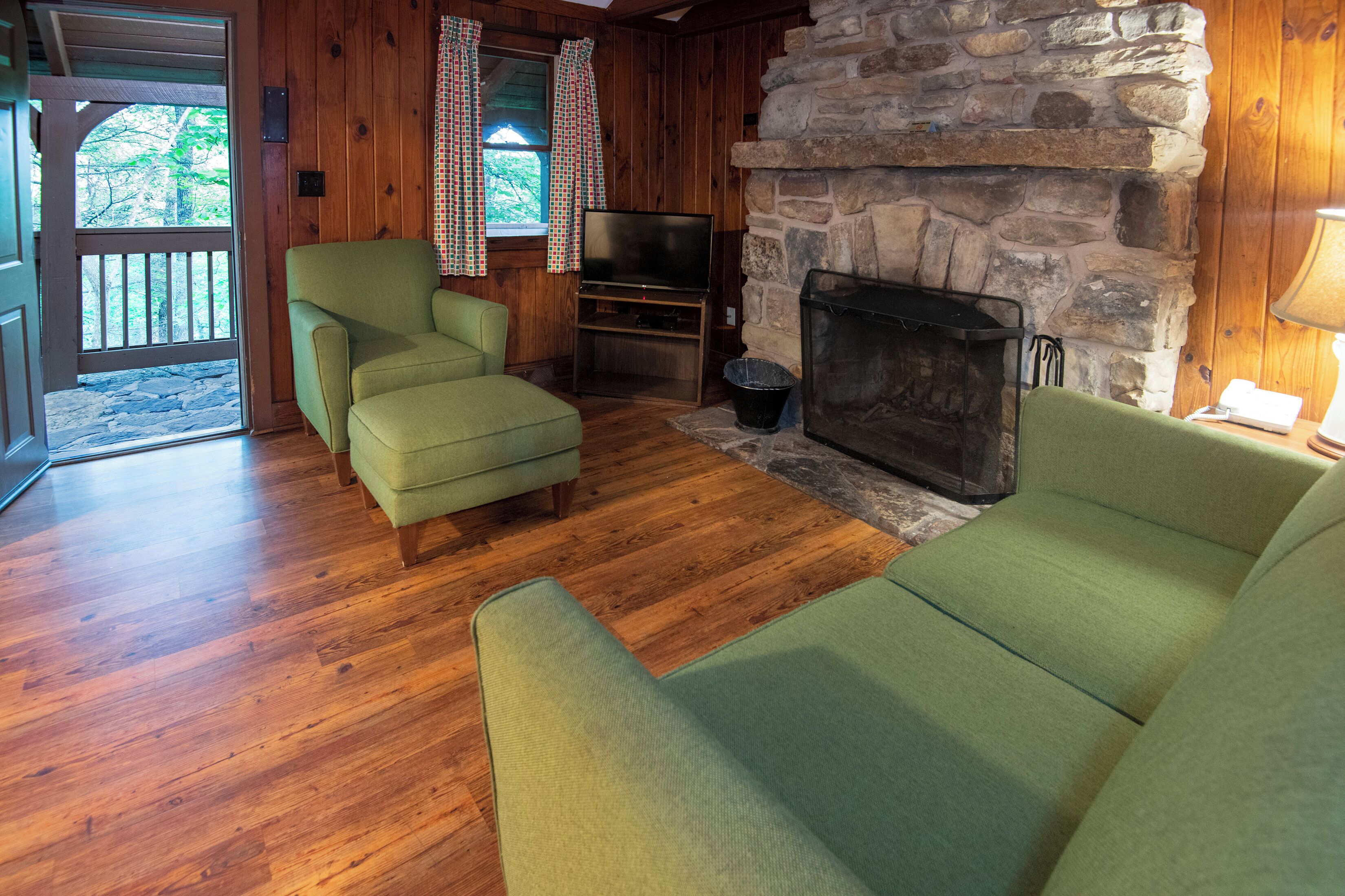 A cozy living room with wooden floors, a stone fireplace, a television, and two green sofas.
