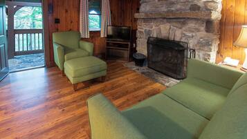 A cozy living room with wooden floors, a stone fireplace, a television, and two green sofas.