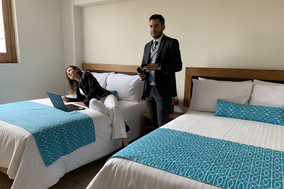 A hotel room with two beds, each with a blue geometric-patterned bedspread. There is a laptop on one bed and a person sitting on the other. A man in a suit stands near the bed on the right.