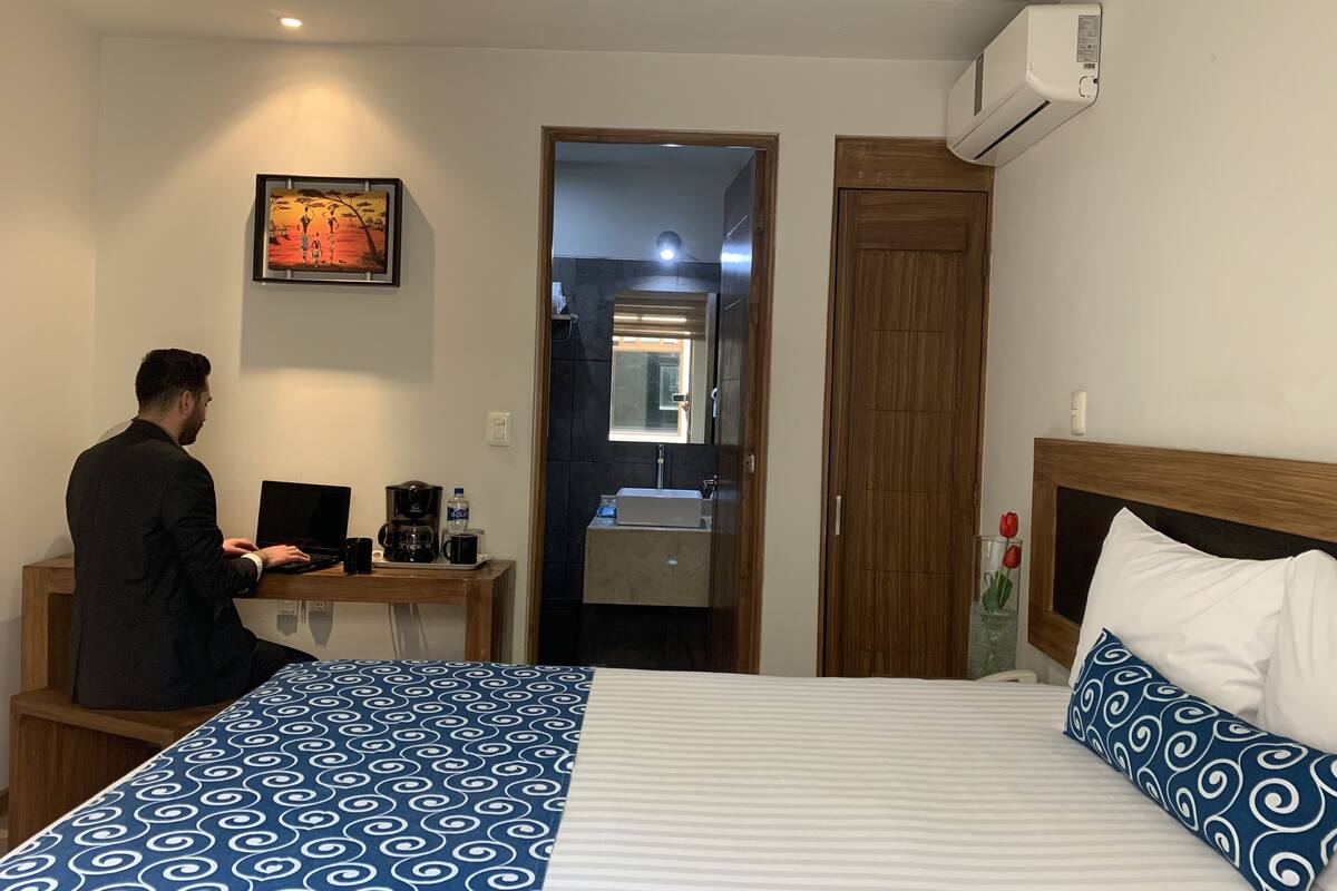A hotel room with a bed, a desk with a laptop, a coffee maker, and a mirror.