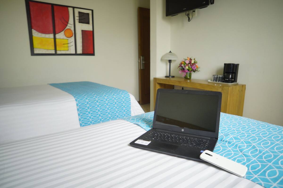 A hotel room with a bed, a desk with a laptop, a coffee maker, and a TV on the wall.