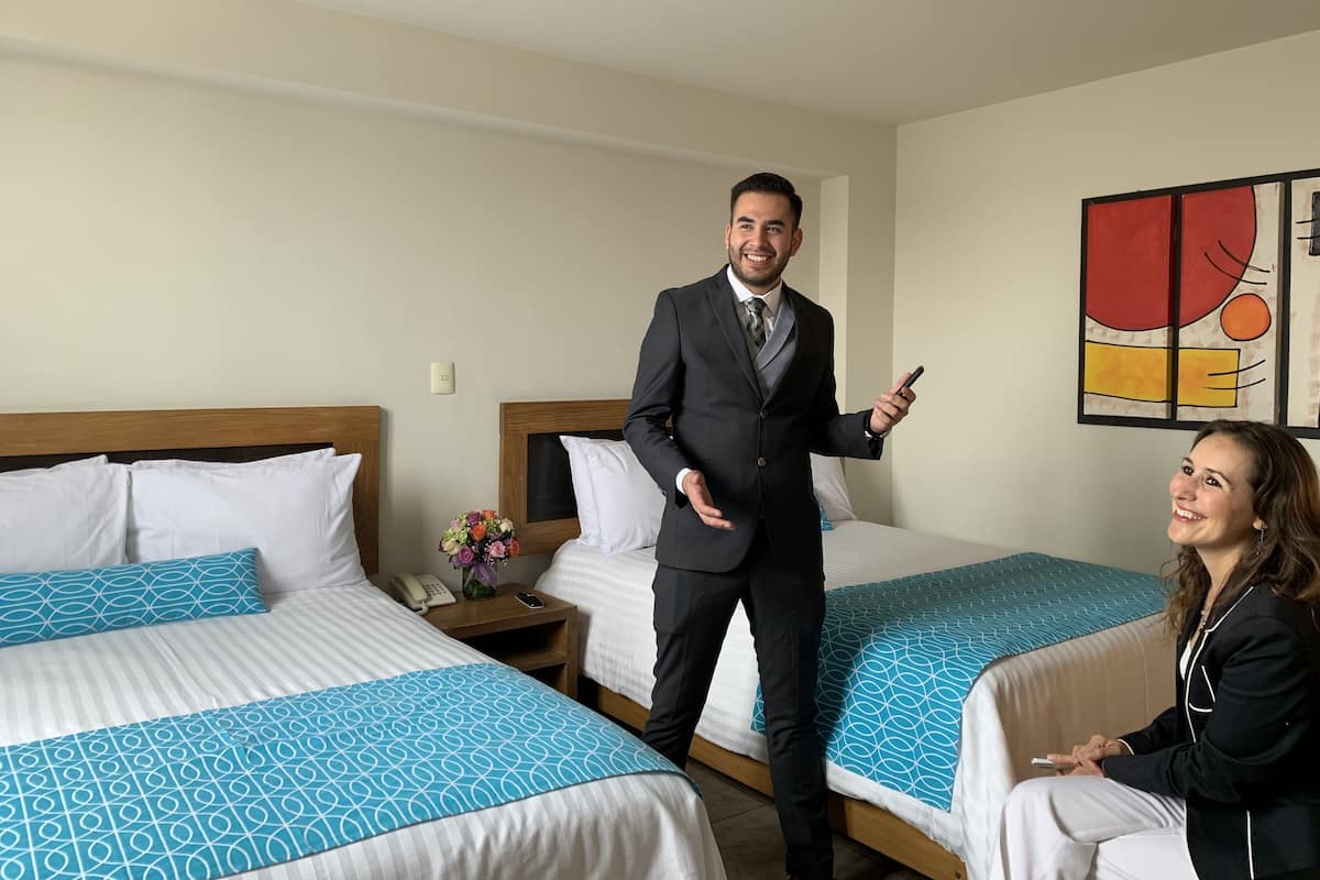 A hotel room with two beds, each with a blue patterned bedspread, a wooden headboard, and white pillows. There is a nightstand with a phone and a vase of flowers. A painting is on the wall.