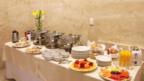 A buffet table with various food items, including fruits, pastries, and beverages, set against a beige wall. - Rymma Hotel (Morelia)