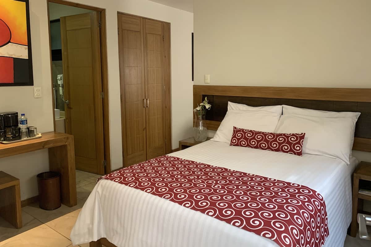 A hotel room with a bed, wooden wardrobe, bedside table, and a small desk with a chair.