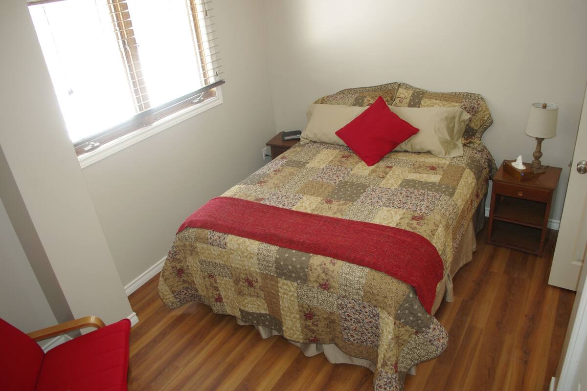 A bedroom with a bed, a nightstand, a lamp, a window with blinds, and a chair.