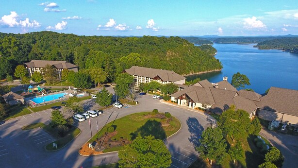 A resort with a swimming pool, parking area, and buildings surrounded by forest and water.