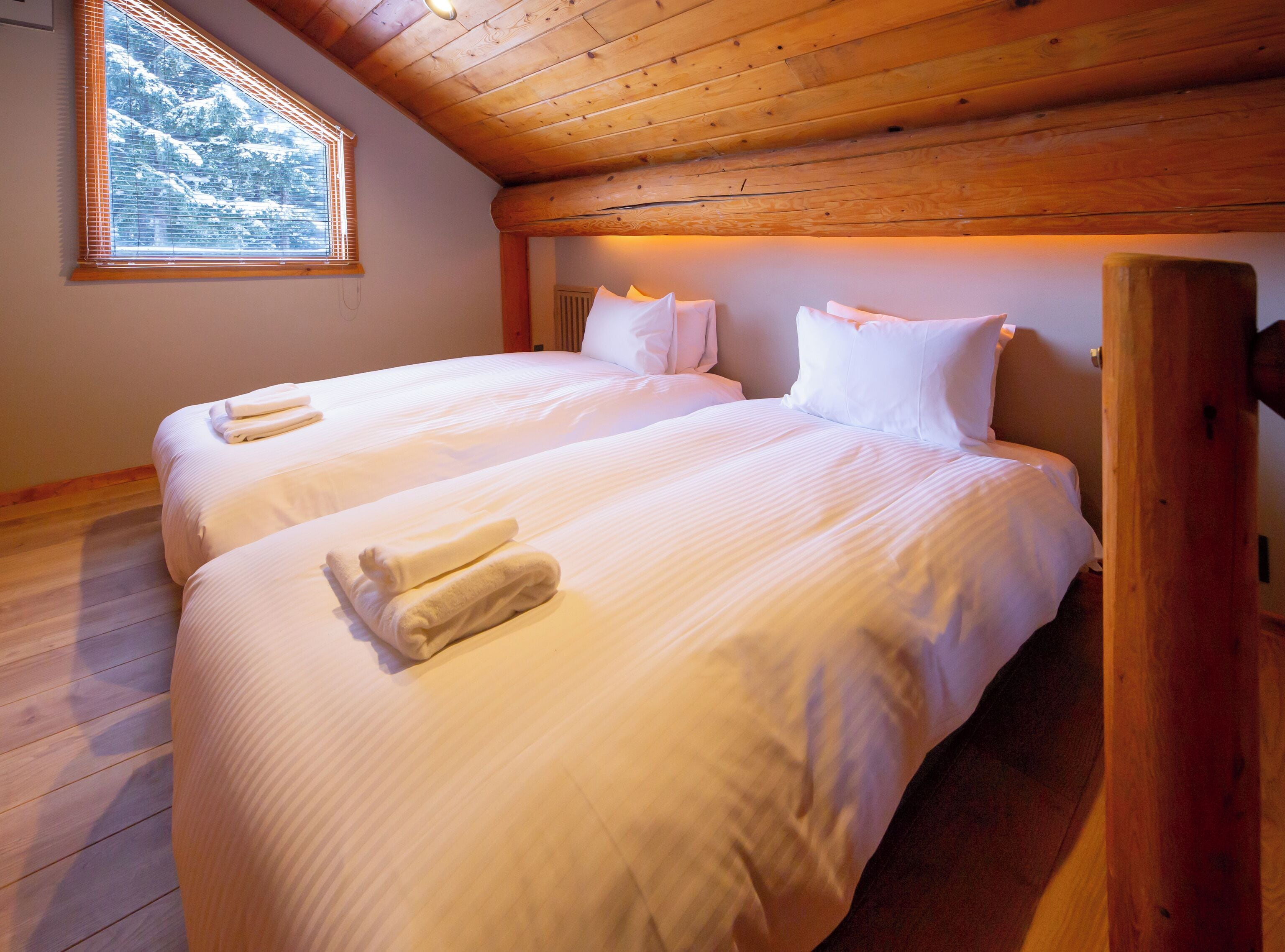 A double bed with white linens and towels, a wooden ceiling, and a window with a view of snow-covered trees.