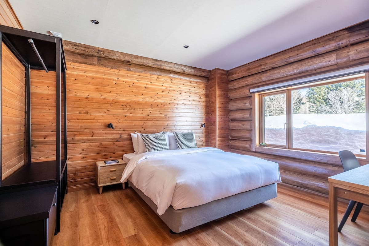 Ein Schlafzimmer mit Wand aus Holzstämmen, einem Bett mit weißer Bettwäsche, einem Nachttisch, einem Schreibtisch und einem Fenster mit Blick auf Bäume.