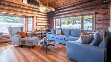 Interior de una cabaña de troncos con un sillón a cuadros, un sofá azul y una mesa de centro redonda.