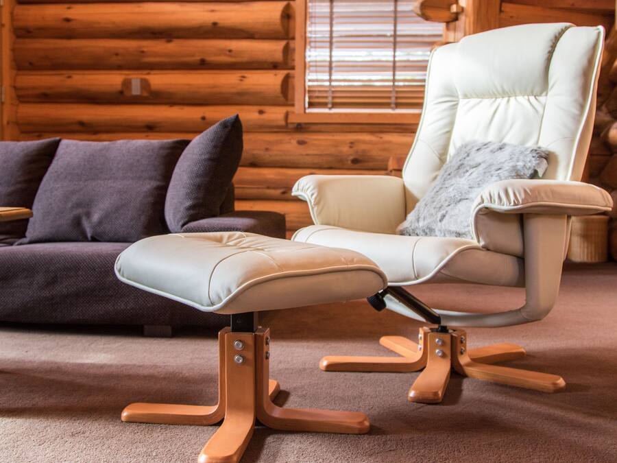 A modern log cabin interior with a beige leather sofa, a light-colored armchair with a fur throw, and a wooden coffee table.