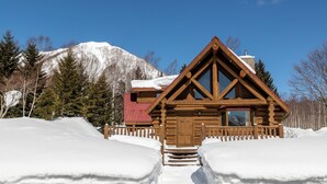 Une cabane en rondins avec un toit à forte pente, un revêtement extérieur en bois et une cheminée. Elle est entourée de neige et offre une vue dégagée sur une montagne en arrière-plan.