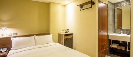 A hotel room with a large bed, a bedside table with a phone, a small table with a kettle, and a bathroom with a sink and mirror.