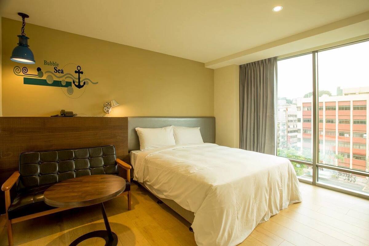 A hotel room with a bed, a chair, a lamp, and a view of the city through large windows.