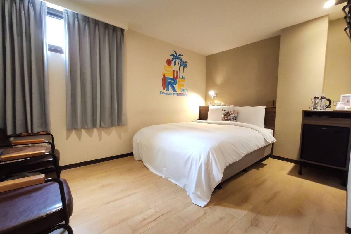 A hotel room with a bed, a desk, a chair, a nightstand, a lamp, and a window with curtains.