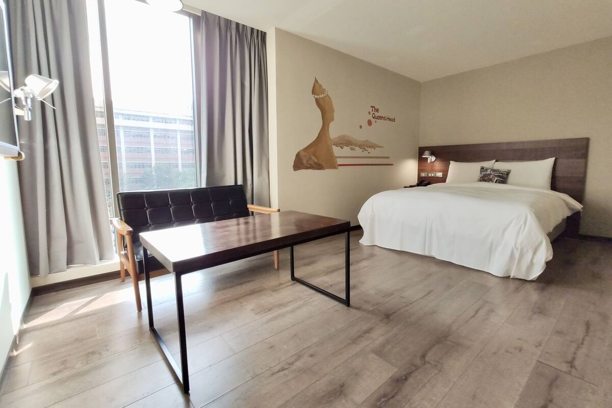 A hotel room with a bed, a desk, a chair, a window with curtains, a wall mural, and a view of buildings outside.