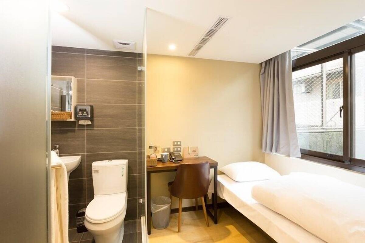 A compact hotel room with a single bed, a small desk, a chair, a toilet, and a sink.