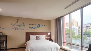 A hotel room with a bed, a nightstand, a lamp, and a large window offering a city view. - Just Live Inn Keelung (Keelung)