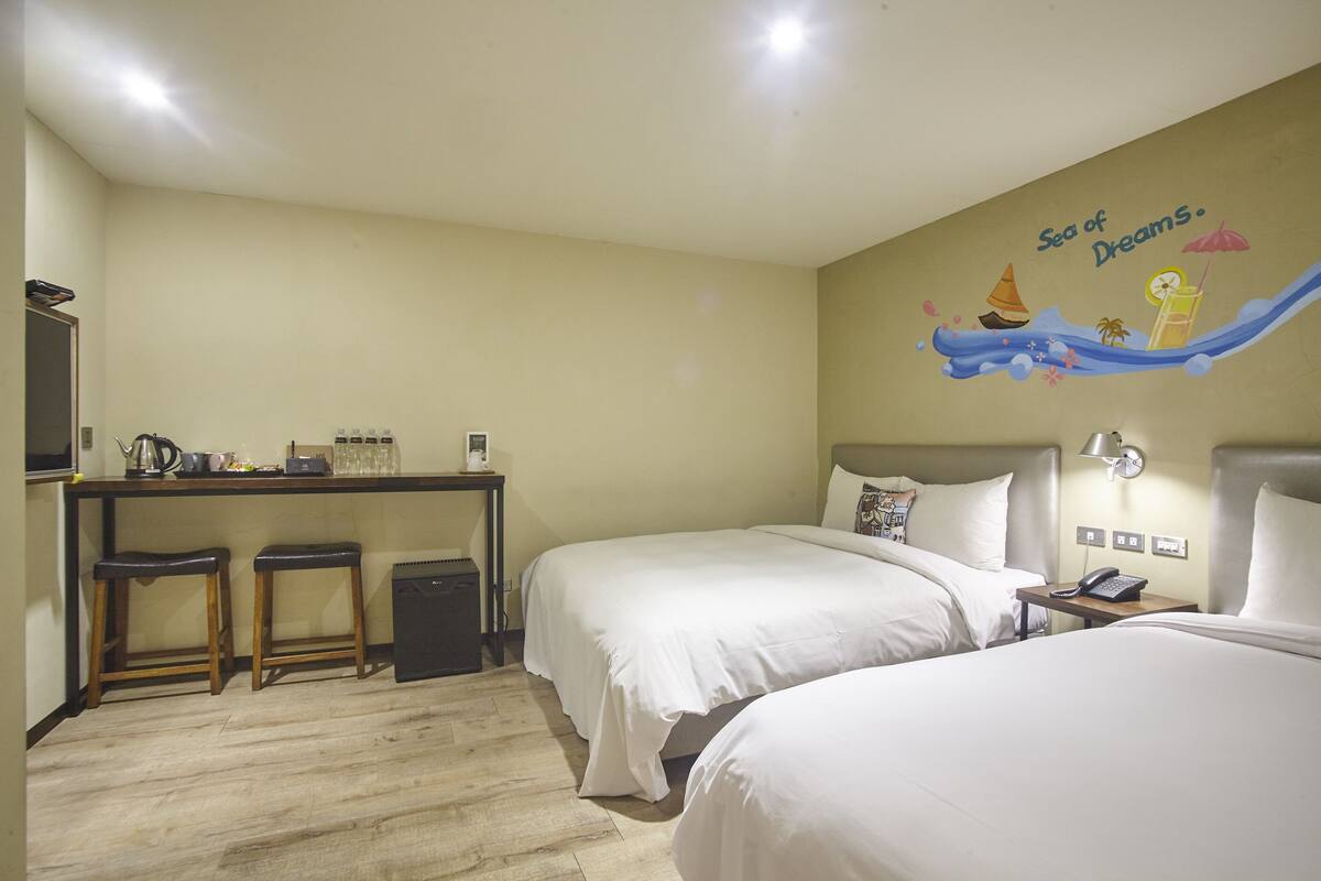 A hotel room with two beds, a desk with stools, a wall mural, and a small table with a phone.