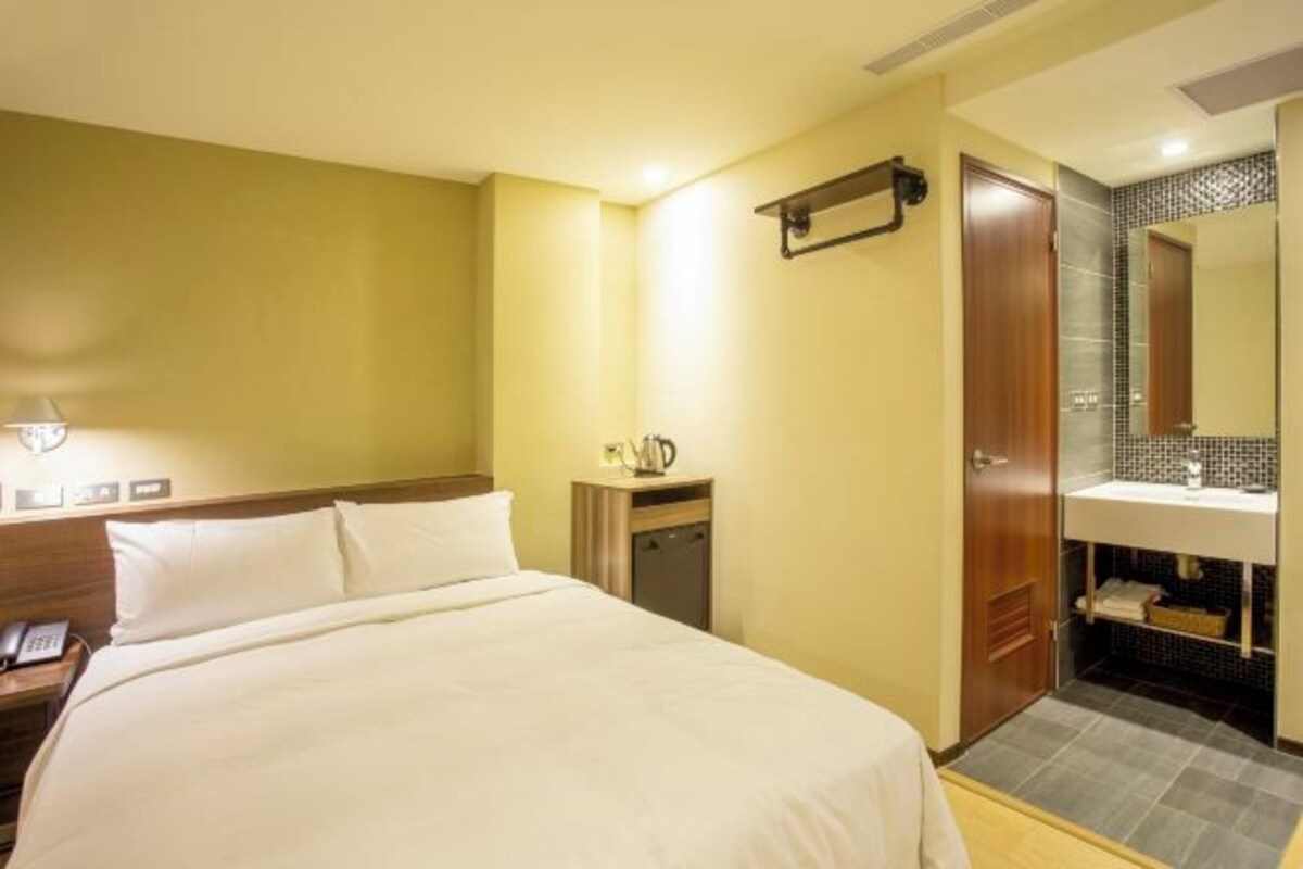 A hotel room with a bed, bedside table with a phone, a small table with a kettle, a mirror above the sink, and a door leading to the bathroom.