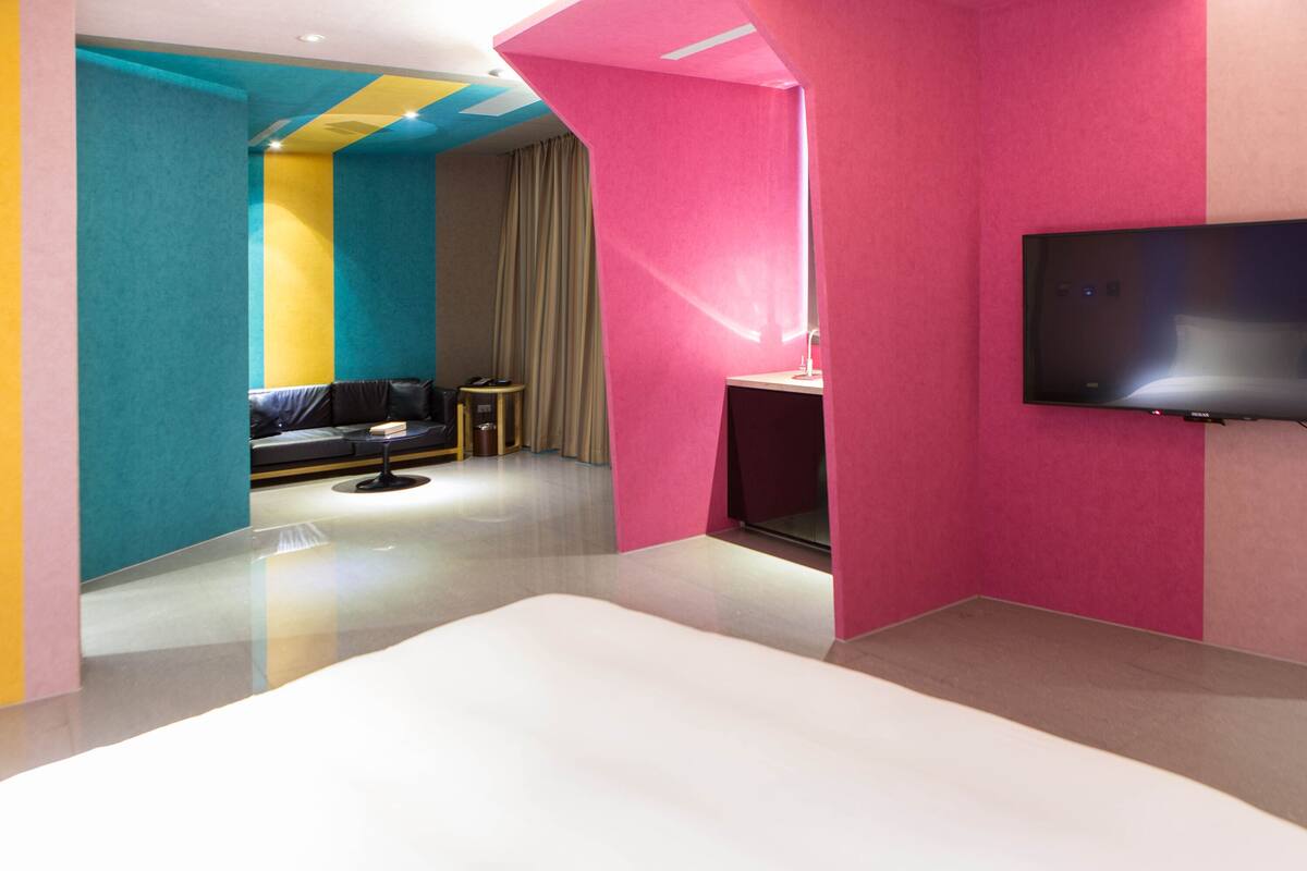 Een moderne hotelkamer met een groot bed, een flatscreen televisie aan de muur, een klein bureau met een lamp, en een zithoek met een bank en een salontafel.