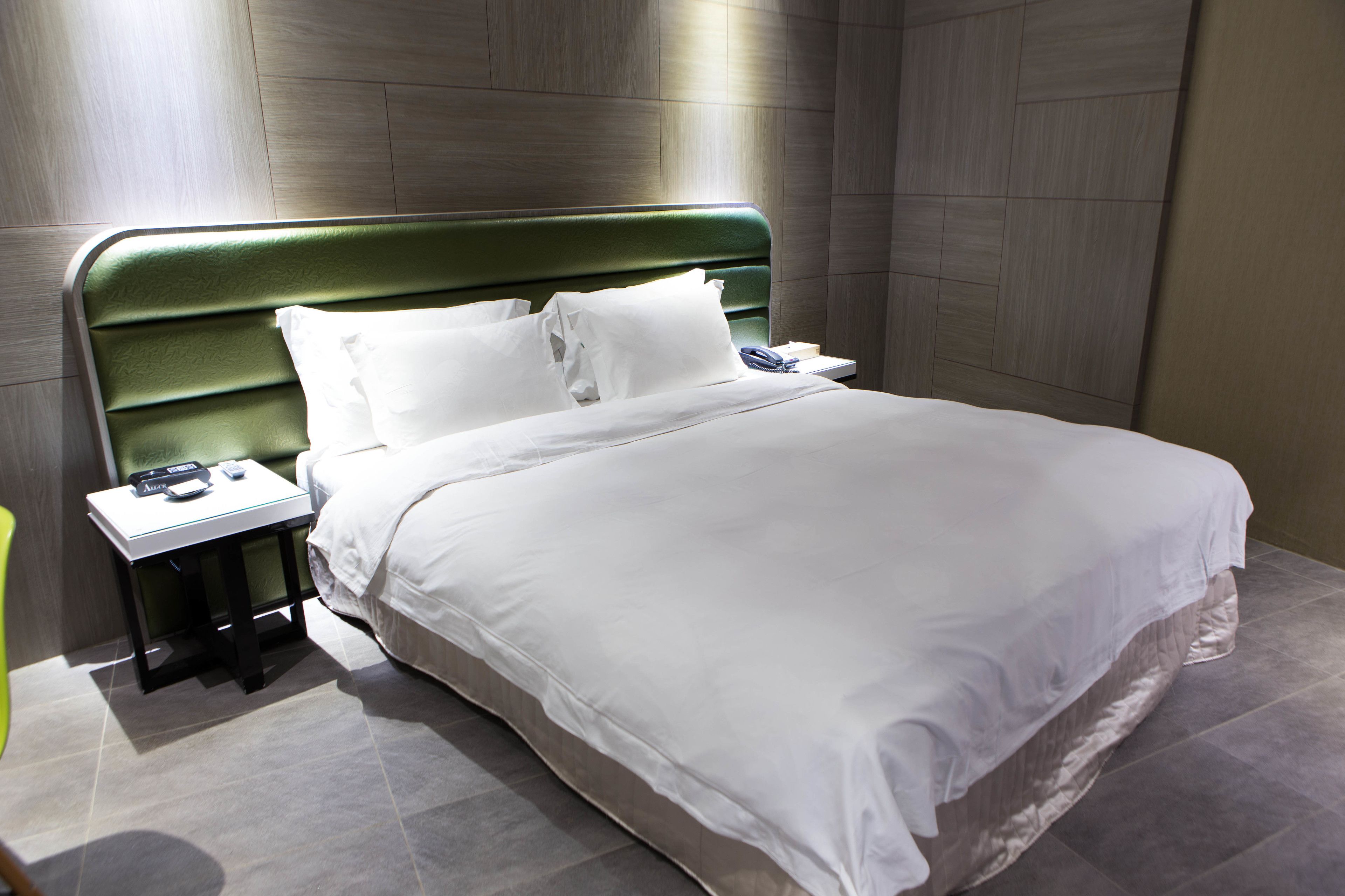 A hotel room with a green upholstered headboard, a white bedspread, and a bedside table with a phone and a lamp.