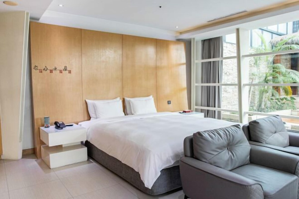 Una habitación de hotel moderna con una cama grande, un sofá gris, un cabecero de madera y un ventanal con vistas a zonas verdes.