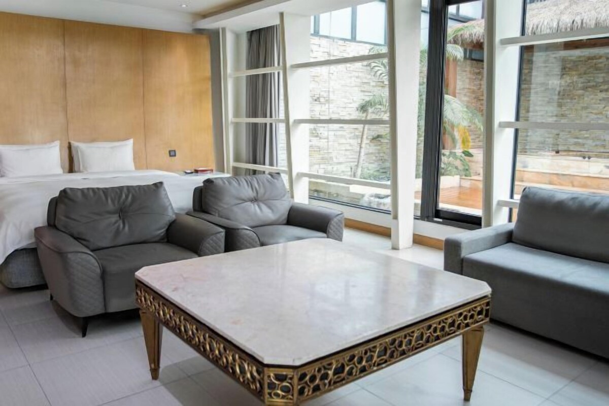 A hotel room with a bed, two grey sofas, a marble coffee table, and large windows offering a view of the outdoors.