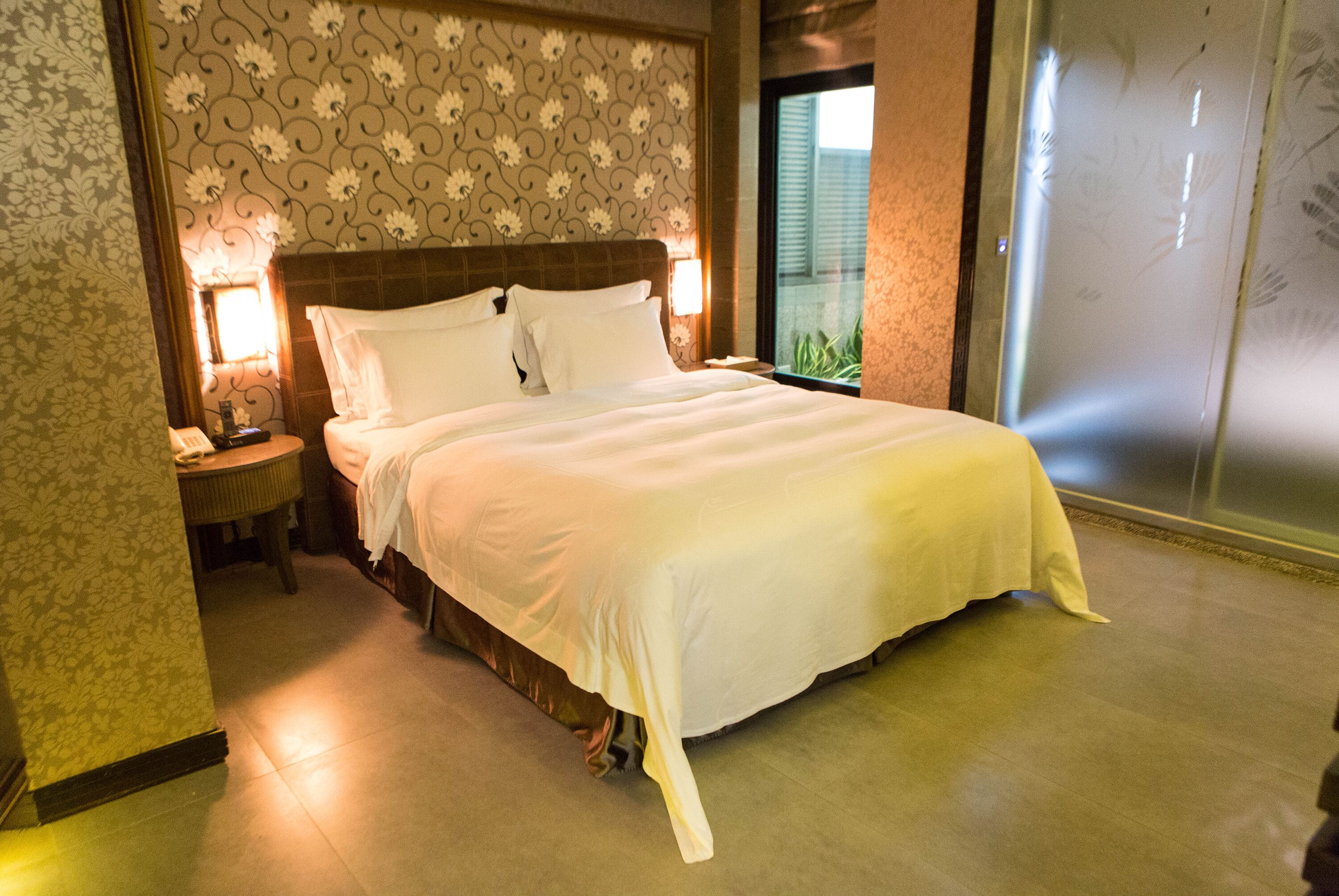 A hotel room with a large bed, a nightstand with a phone, and a window with a view of greenery.