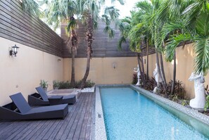 Un área de piscina con sillones de descanso, palmeras y estatuas decorativas.