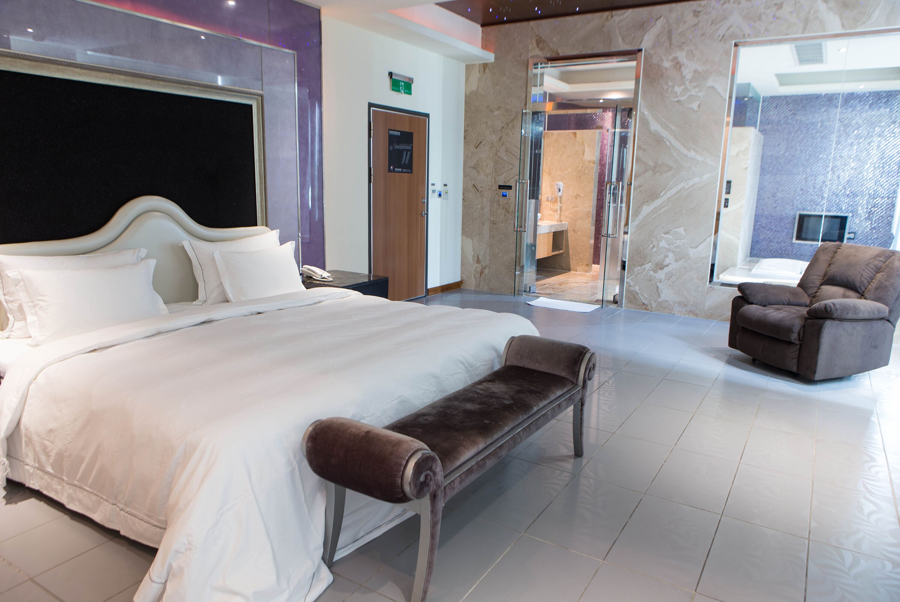 A modern hotel room with a large bed, a bench, a chair, and a bathroom visible through a glass door.