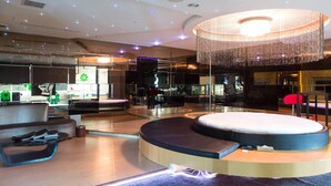 A modern hotel lobby with a circular seating area, a large chandelier, and a glass partition with a green logo. - All-Ur Boutique Motel - Ping-Tung Branch (Pingtung)