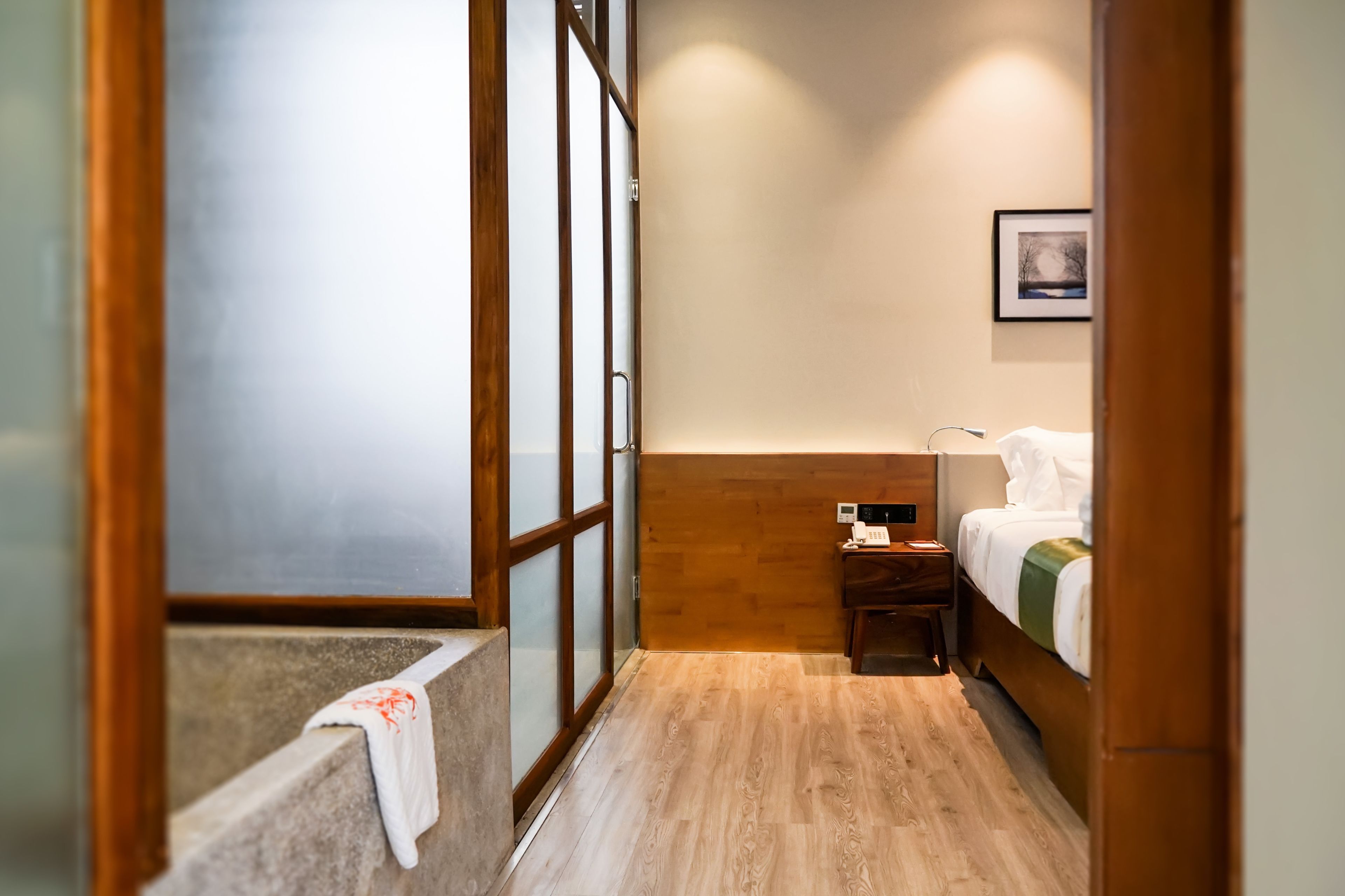 Habitación de hotel con piso de madera, una cama con colcha verde y blanca, una mesita de noche con lámpara y un cuadro en la pared.