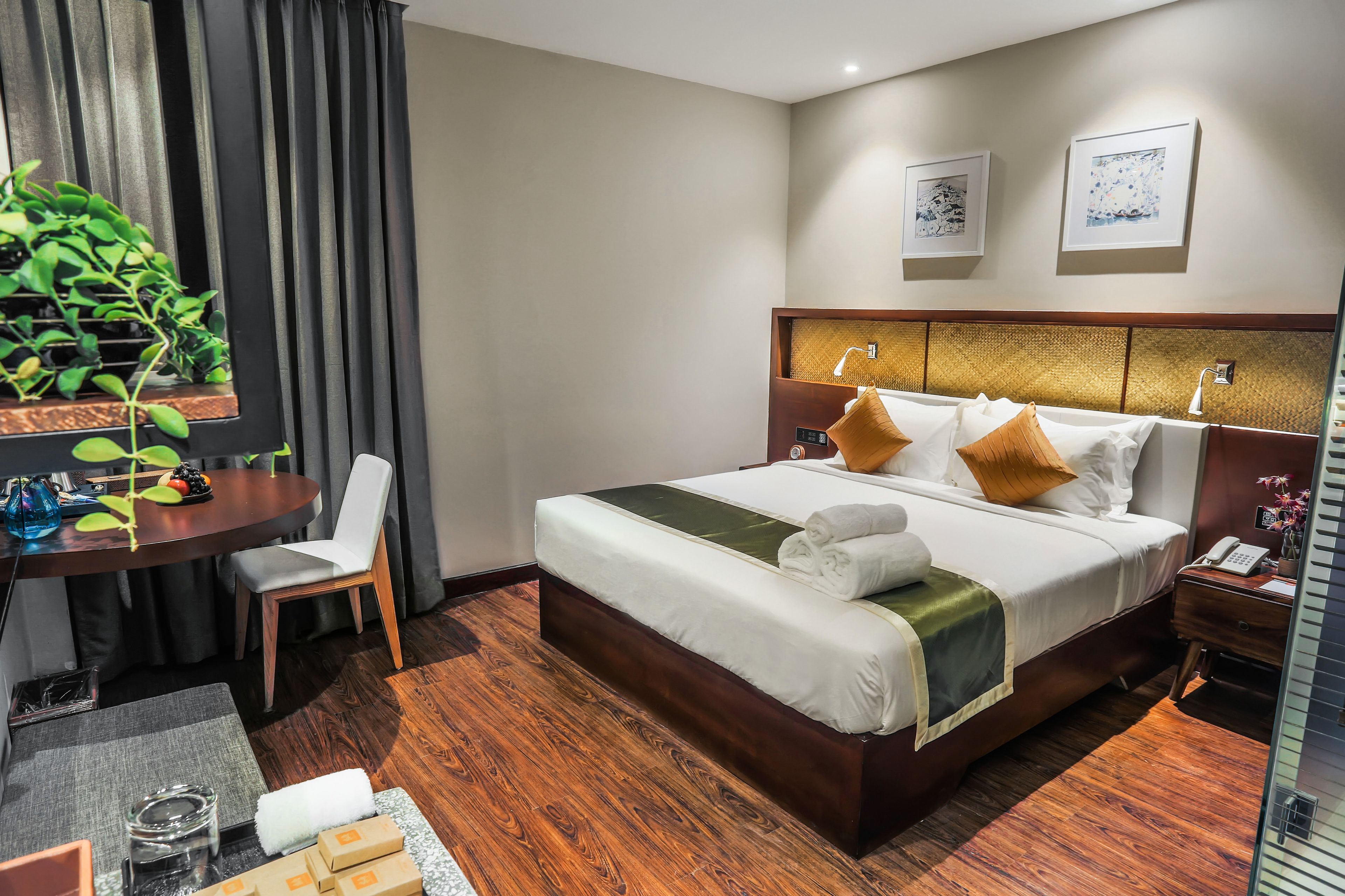 A hotel room with a large bed, a desk, a chair, a small table with a plant, and a bedside table with a lamp.