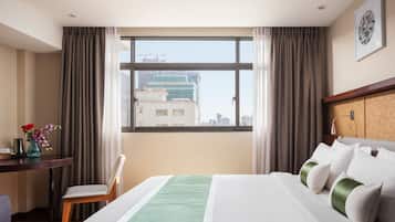A hotel room with a large window, a bed with white and green bedding, a desk with a vase of flowers, and a chair.