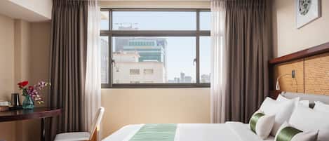 Habitación de hotel con un ventanal grande, cama con ropa de cama blanca y verde, escritorio con un florero y una silla.
