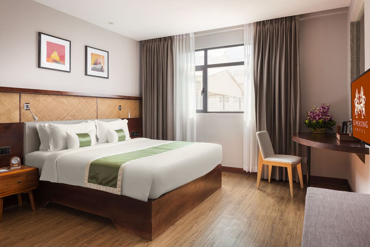 A hotel room with a large bed, a wooden nightstand, a desk with a chair, a window with curtains, and framed artwork on the wall.